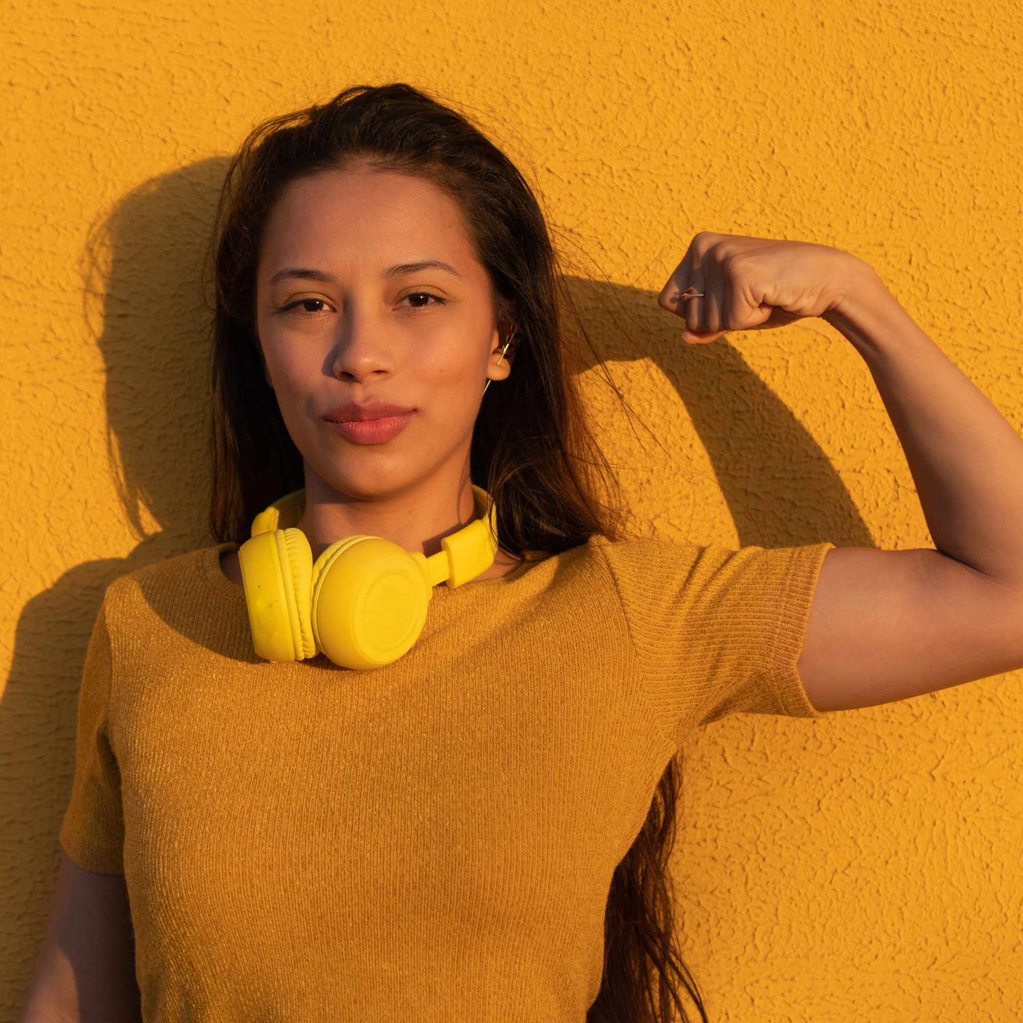 Portrait d’une femme devant un mur jaune, portant un t-shirt jaune et un casque audio autour du cou, levant le bras en signe de force et de confiance.