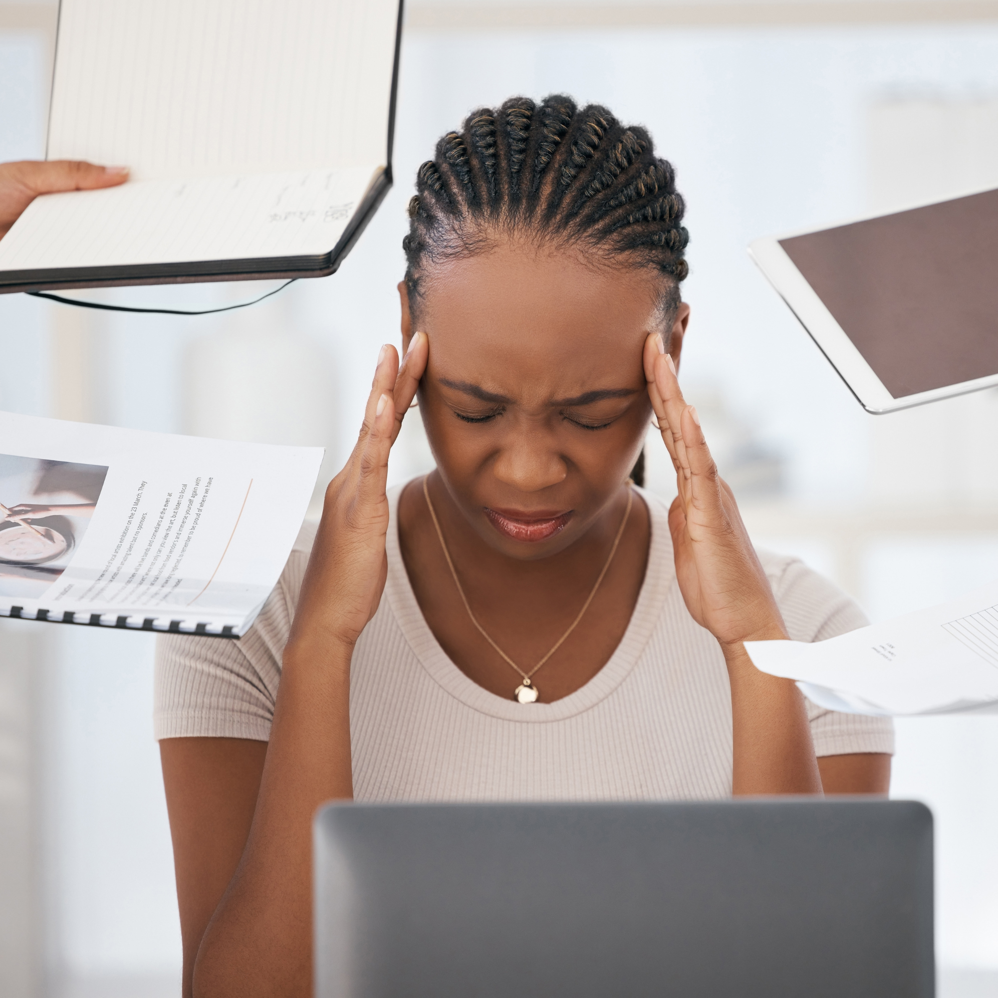 Les perfectionnements : Femme assise devant un ordinateur, les yeux fermés et les mains sur les tempes, semblant stressée, avec des documents et objets en suspension autour d’elle illustrant une surcharge mentale.